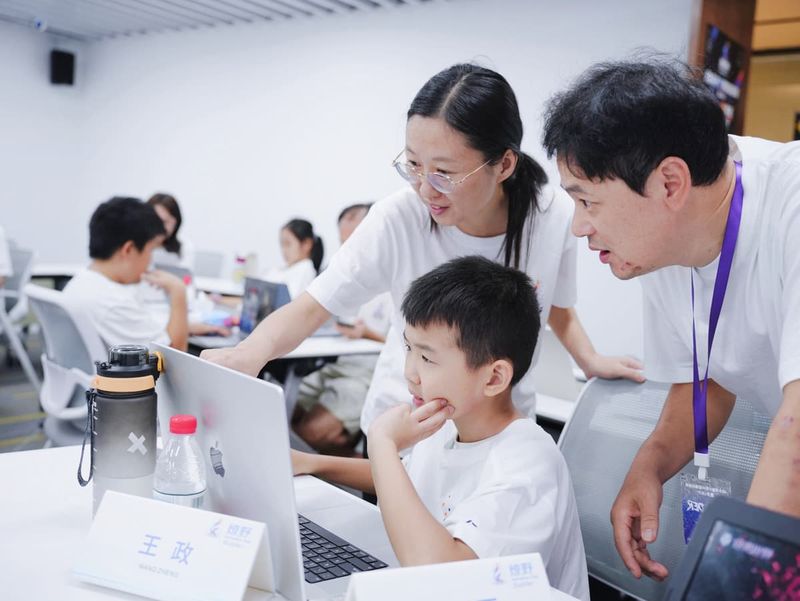 燎野 48 小时青少年 AI 黑客松现场：一个孩子托着下巴盯着电脑，两位大人凑过来一起看屏幕
